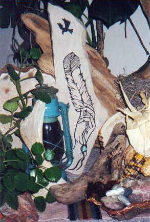 Long irregularly shaped flat sandstone, 
leaning against driftwood, surrounded by 
smaller driftwood, stones, dark spackled 
yellow maize and kerosene lamp.  
Graph carved into the slab shows a slender 
female hand holding up a dark tipped eagle 
feather to an Eagle shade soaring above 
looking down. The shaft of the feather 
decorated with dangling strings.