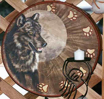 Round drum painted. 
A dark colored Wolf stands left in front of 
a radiating halo around its head and breast. 
Wolf looks East with pointed attentive ears 
and open mouth, showing its white teeth. 
Seven wolf paw- or footprints run along the 
drum border from center top to left lower left. 
Each heart shaped metacarpal pad with four 
digital pads and claw imprints beneath.